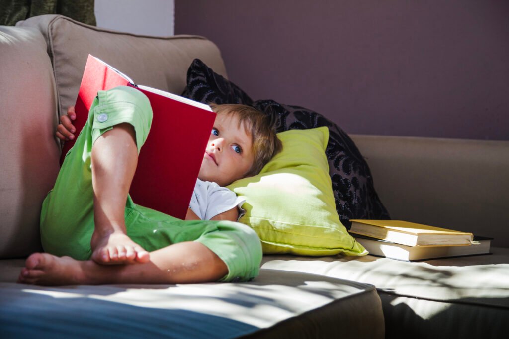 Menino deitado no sofá fazendo a leitura de um livro.