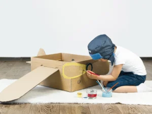 Suporte Pedagógico: menino pitando uma caixa de papelão transformada em um avião.