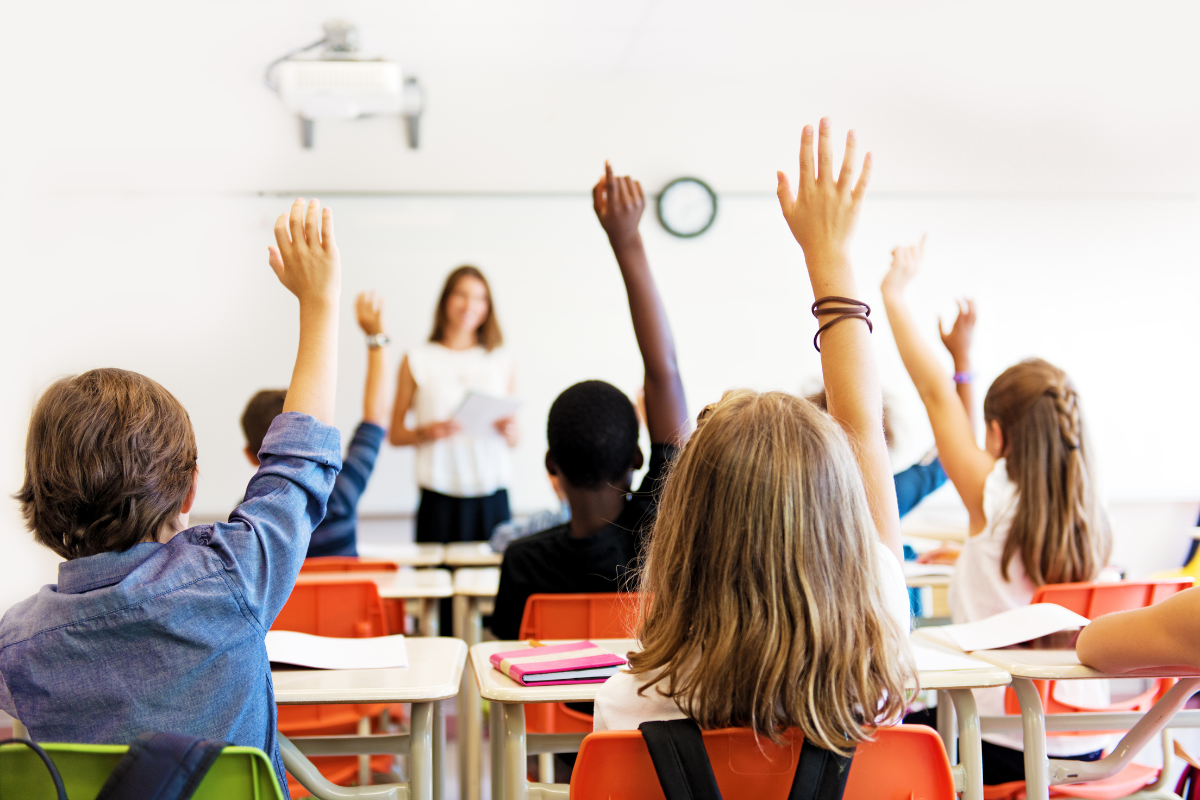 Alunos com as mãos levantadas e a professora olhando para eles.